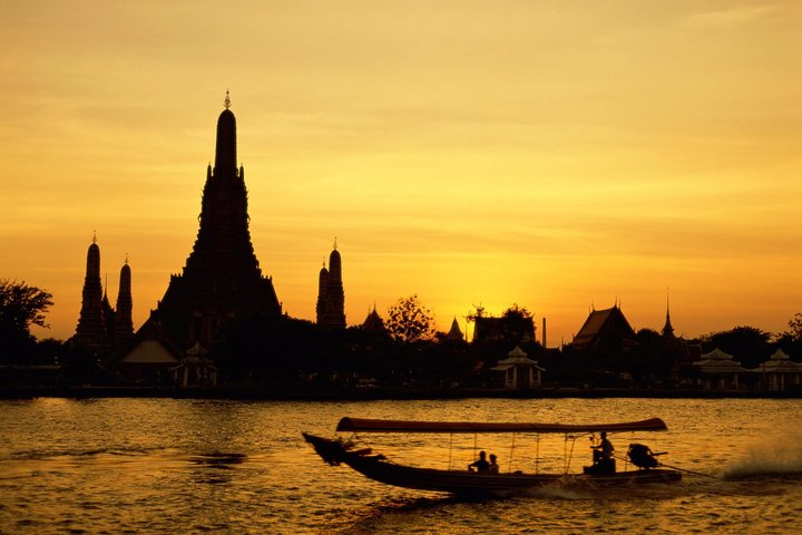 Golden hues reflect off the water as a boat glides past ancient temple silhouettes inviting travelers to uncover the rich history of Ayutthaya's majestic ruins.