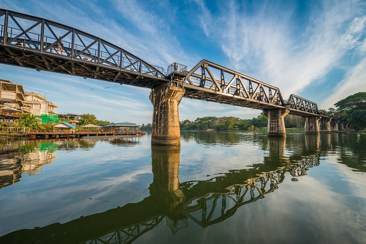 Experience the charm of Kanchanaburi as the iconic Bridge over River Kwai stands tall against the serene water reflecting the rich history and scenic beauty of this unforgettable destination.