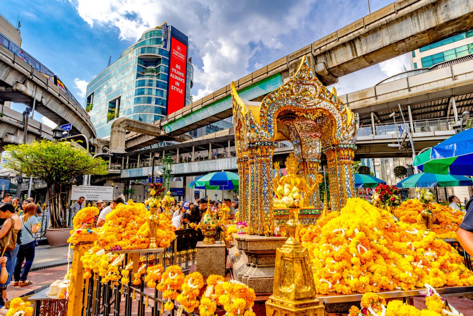 Experience the serenity of the Four-Faced Buddha with vibrant floral offerings surrounding a stunning shrine creating a unique space for reflection and wish-making in bustling Bangkok.