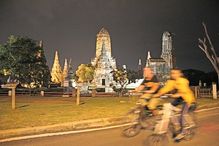 Discover the charm of Ayutthaya as you cycle through illuminated temple ruins at sunset enjoying a unique perspective on this historic site while soaking in the serene night scenery.