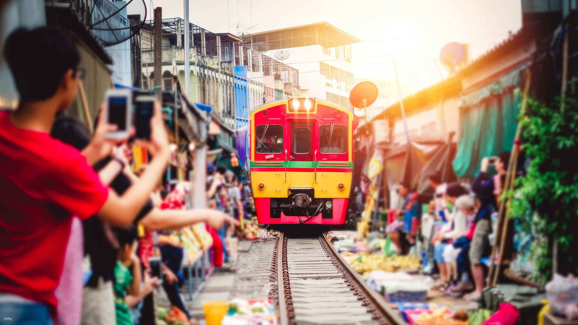 Experience the unique charm of Thailand as a train passes through the bustling market surrounded by colorful stalls and eager onlookers capturing a moment of local life.