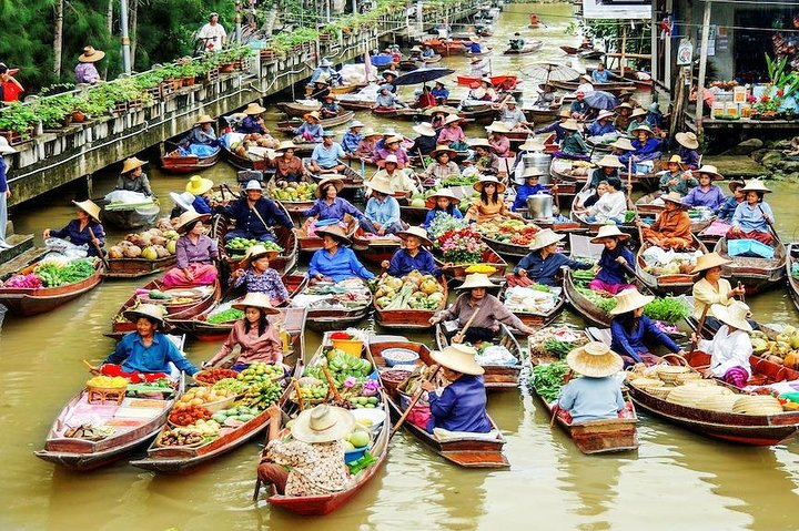 Colorful wooden boats filled with fresh produce glide through serene waters showcasing the timeless charm of local life at Thaka Floating Market. Experience authentic Thai culture amidst the bustling scene.